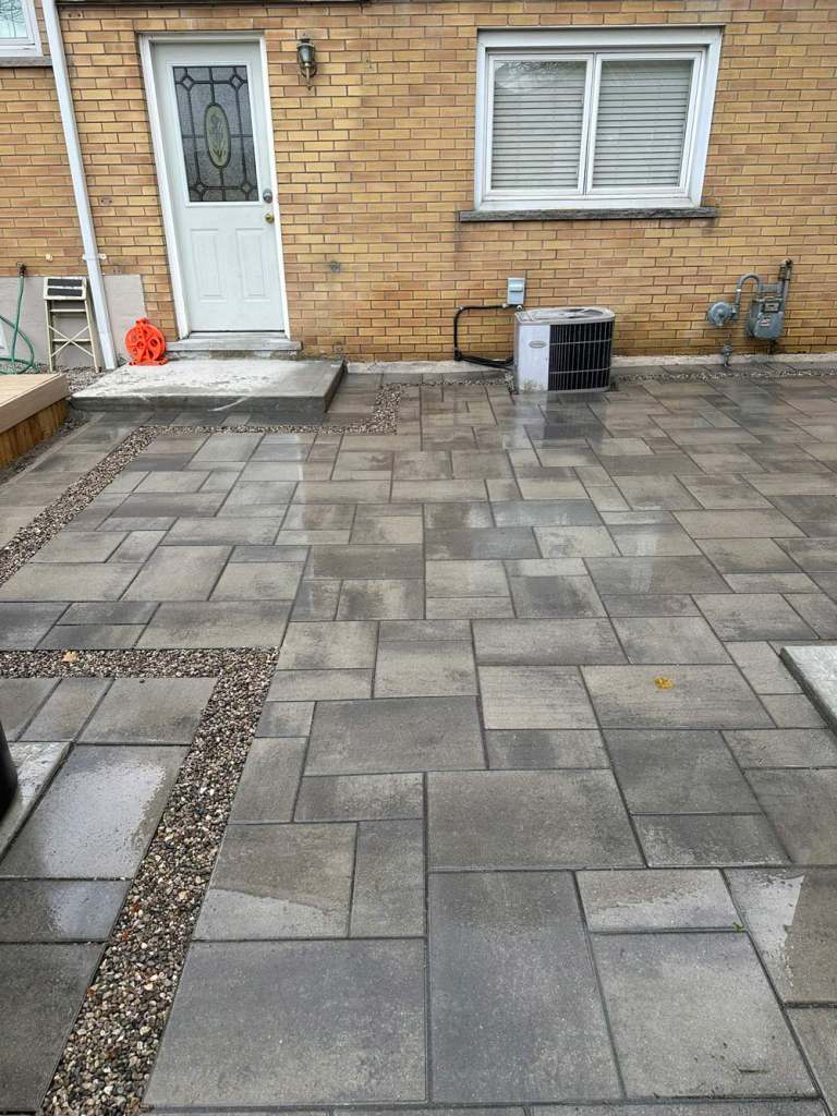 Grey interlock patio at a back door with a pea gravel inlay strip, yellow brick house