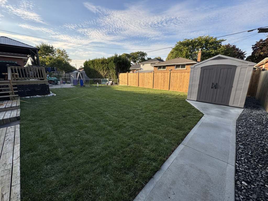 Lumen Landscaping | When the Water Comes From Next Door New sod lawn and a concrete walkway to a shed edged with dark river rock in a Hamilton Mountain backyard