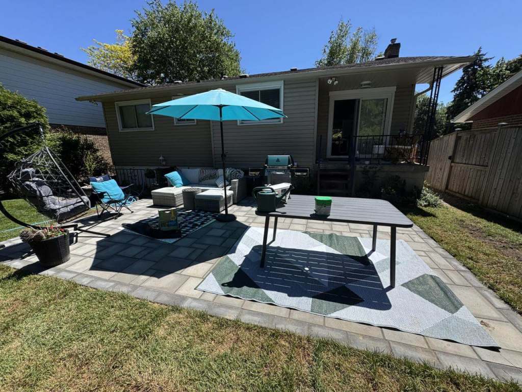 Lumen Landscaping | How to Compare Landscaping Quotes in Hamilton: 10 Questions to Ask Grey interlock patio behind a house with an outdoor sofa, dining table and a teal umbrella