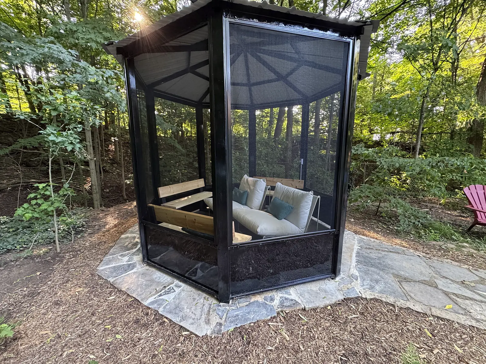 Screened gazebo with wood bench seating set on a flagstone pad in a wooded backyard