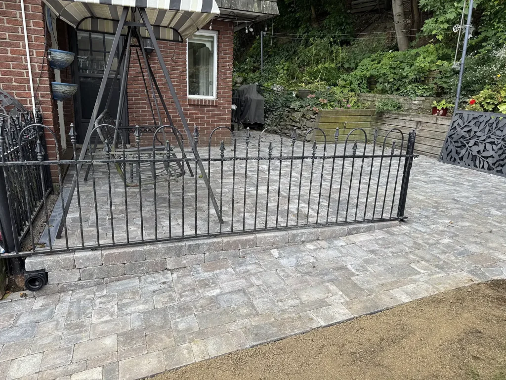 Lumen Landscaping | Turning a Sloped Escarpment Yard Into Usable Space Grey interlock patio with a black metal fence and a raised garden wall behind it