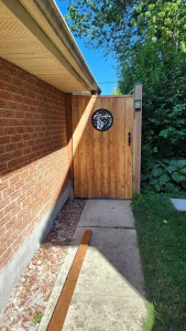 Lumen Landscaping | Fences and Gates Wood side gate with a round decorative metal inset between a brick house and a fence